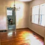 An empty room with wooden floors, a chandelier, and a person sitting in a chair visible through an open doorway.