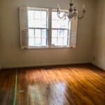 Empty room with hardwood floors, two windows with shutters, and an unlit chandelier hanging from the ceiling.