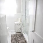 Bright bathroom with white walls, a glass shower, a small rustic cabinet, a fluffy gray rug, and a window with sheer curtains.