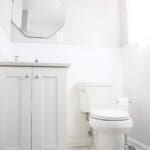 Minimalist white bathroom with octagon mirror, vanity cabinet, toilet, and a window with sheer curtain, illuminated by natural light.