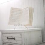 Open book with sheet music and text on a small white cabinet, set in a bright room with white paneled walls.