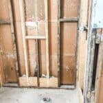Exposed wall studs and plumbing in a bathroom undergoing renovation, with a drain opening visible on the unfinished floor.