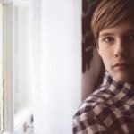Boy wearing a plaid shirt, standing by a window with white curtains, looking directly at the camera.