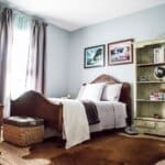Cozy bedroom with a wooden bed, gray bedding, wicker basket, bookshelf, and framed art above the bed on gray walls.