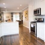 Modern kitchen with white cabinets, stainless steel appliances, hardwood floors, and an island. Living room with patterned curtains visible in the background.