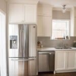 Modern kitchen with white cabinets, stainless steel refrigerator, dishwasher, and sink under a window with natural light.