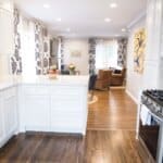 Modern kitchen with white cabinets and countertops, opening into a living room with hardwood floors, patterned curtains, and cozy seating.