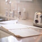 Open spiral notebook on a kitchen counter beside a stainless steel toaster and sink with a blurred background.