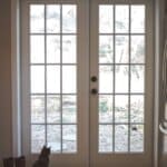 Cat sitting by French doors, looking outside. Boots and patterned rug are by the doors, with natural light illuminating the space.