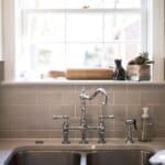 Modern kitchen sink with chrome faucet, soap dispenser, and a window above with a cutting board, black cat statue, and plants.