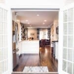 Open double doors reveal a bright kitchen with white cabinets, stainless steel appliances, and an adjoining living area.