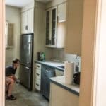 A person crouches by the refrigerator in a small kitchen with white cabinets, a dishwasher, and a roll of paper towels on the counter.