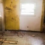 A room under renovation with a yellow and white wall, a single window, exposed wiring, and a partially removed floor section.