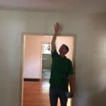 A man in a green shirt reaches up to touch the ceiling inside a painted room with hardwood floors and adjacent room entry.