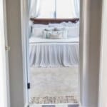 Hallway with black and white checkered floor leading to a bedroom decorated with white linens and multiple pillows.