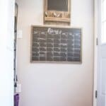 Wall-mounted calendar and chalkboard with a "Remember to buy" list. Below are two pet bowls on the floor near a door.