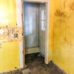 A doorway leading to a small hallway with damaged yellow walls, exposed wires, and a dirty floor, indicating renovation work.
