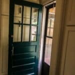 Green wooden door with glass panes open to porch, sunlight entering the dim-lit room through the screen door.