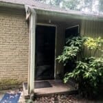 A small house entry with a black screen door, broken gutter, and leafy plants on the right, surrounded by greenery and fallen leaves.