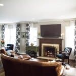 A cozy living room with a fireplace, TV above it, leather sofa, patterned curtains, and an adjacent kitchen with a small dining area.