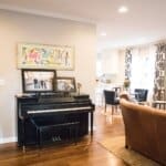 Open-concept living area with a piano and family photos, wooden floors, leather sofa, and kitchen with black-and-white curtains.