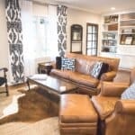 A cozy living room with leather furniture, a wooden coffee table, a cowhide rug, built-in shelves, and patterned curtains.