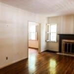 Empty room with hardwood floors, a fireplace, and two windows with shutters. Adjacent room visible through an open doorway.
