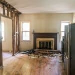 Renovation in progress with torn-down walls, exposed beams, scattered debris, and a fireplace between two windows.