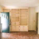Empty living room with open blue door, built-in cream-colored shelves, hardwood floor, and stainless steel refrigerator.