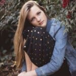 A young woman with long blonde hair, wearing a denim jacket, sits surrounded by foliage with red berries, sunlight behind her.