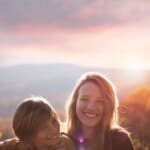 Two smiling children sit closely together outdoors at sunset, with mountains and a colorful sky in the background.