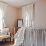 Bright bedroom with white curtains, a wooden bed, and an antique sofa in front of windows. Beige walls and wooden floor.