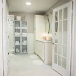 Bright bathroom with a glass cabinet filled with white towels, a double sink with mirrors, and a walk-in shower in the background.