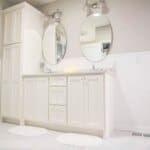 Modern bathroom with white double vanity, oval mirrors, and wall-mounted lights. Linen closet with neatly folded towels is on the left.