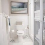 Clean white bathroom with a toilet, towel rack, and storage shelves. A beach painting hangs above the wainscoting.