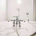 White tiled shower with a vintage chrome showerhead, two other fixtures, and a long coiled hose.