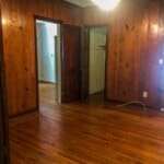 Interior of a room with wooden panel walls, hardwood flooring, a doorway leading to another room, and a ceiling light.