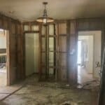 Interior room under renovation with exposed wooden framing, debris on the floor, and light fixtures hanging from the ceiling.