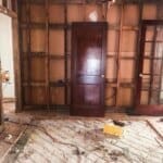 A room under renovation with exposed wall studs, wooden doors, debris on the floor, and a hanging light fixture.