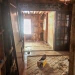 A partially renovated room with exposed beams, wooden floors, and debris scattered around, illuminated by natural light.