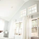 A bright bedroom with vaulted ceilings, white walls, large windows with curtains, a bed, a wicker basket, and a wooden desk.
