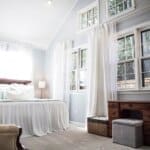 Bright bedroom with large windows, white curtains, a cushioned bed with a wooden headboard, and a wooden desk with a gray ottoman.