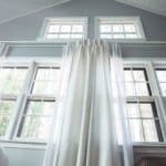 Large windows with white curtains fill a high-ceiling room, letting in natural light and showcasing an outdoor view.