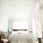 Bright, airy bedroom with a large bed, white bedding, fluffy pillows, and a window with sheer curtains, flanked by two table lamps.