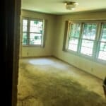 Empty room with beige carpet, large windows letting in natural light, and a ceiling light fixture.