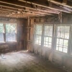 Partially demolished room with exposed wooden beams, insulation removed, and large windows letting in natural light from outside.