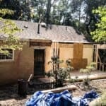 Rear view of a house under renovation with a partially constructed wooden addition and blue tarps on the ground.
