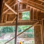 Partially constructed wooden house interior with open windows, showing a person working outside surrounded by trees and greenery.