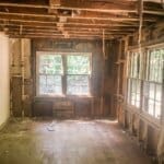 A partially gutted room with exposed wooden framing, missing drywall, and numerous windows allowing natural light.